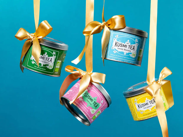 Cztery kolorowe puszki herbaty Kusmi, każda ozdobiona złotym kokardą i z unikalnymi etykietami dla różnych odmian herbaty.
